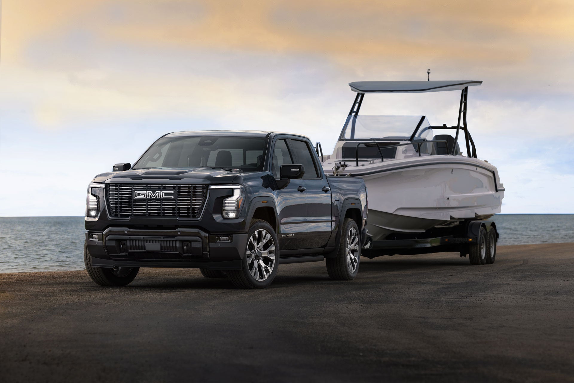A black GMC Denali truck towing a white motorboat on a trailer, parked on a boat ramp next to a lake.